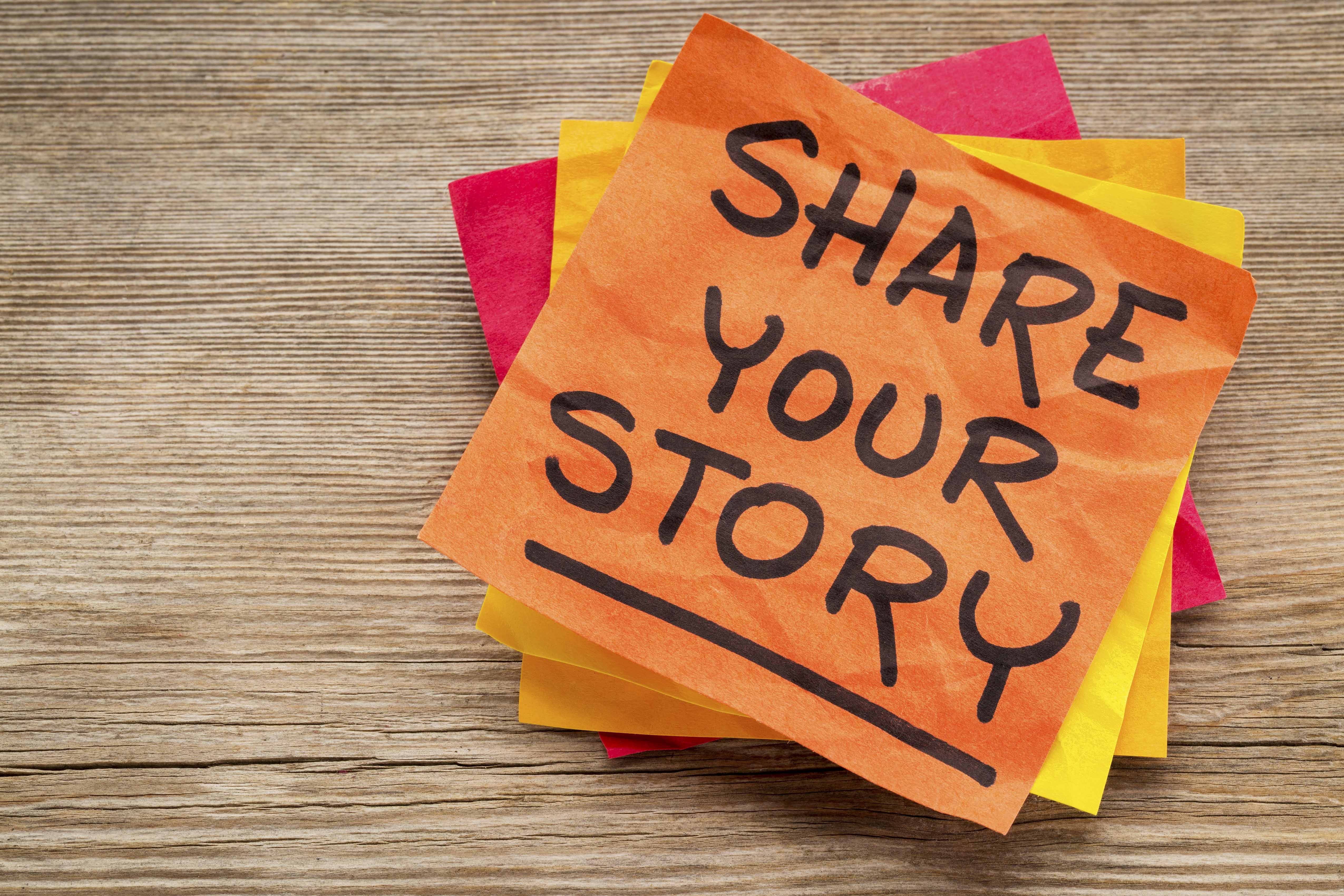 Note that says share your story suggestion on a sticky note against grained wood.